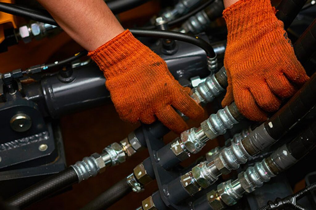 建設現場で作業員が油圧ショベルの油圧ホースを交換・圧着し、作動油を補充している様子